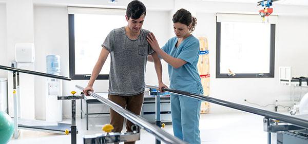 Maennlicher Patient bei der Physiotherapie geht mit Hilfe der Barren und des Therapeuten neben ihm Männlicher Patient bei der Physiotherapie geht mit Hilfe der Barren und des Therapeuten neben ihm