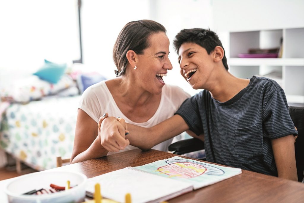 Teenager mit Behinderung und seine Mutter sitzen am Tisch und lachen