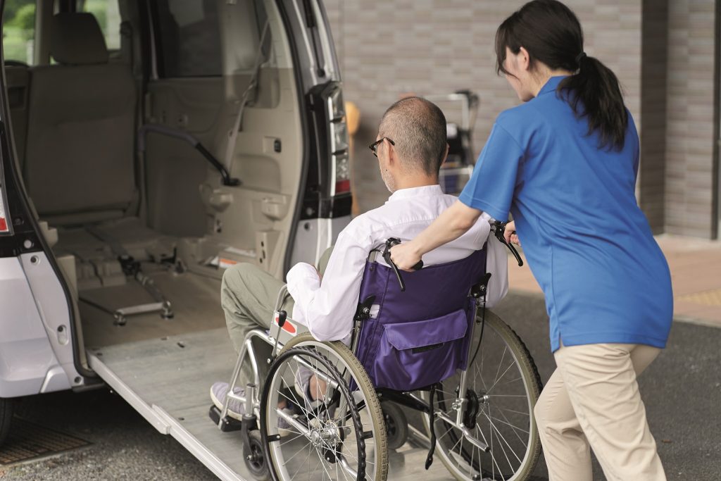 Eine Pflegekraft schiebt einen Herrn im Rollstuhl über eine Rampe in ein Fahrzeug