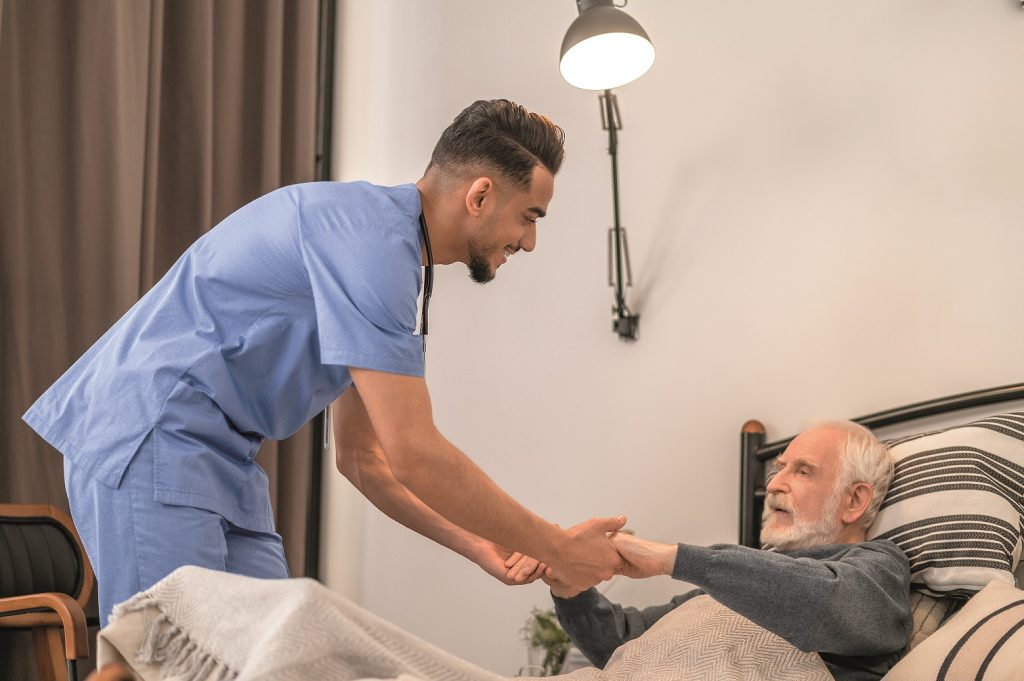 Männliche Pflegekraft hilf älterem Mann beim Aufrichten im Bett Männliche Pflegekraft hilf älterem Mann beim Aufrichten im Bett